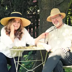 Para z kapeluszami siedzi przy stoliku w ogrodzie. Kobieta, jakby zainspirowana twórczością Daukszewicza, bawi się jego krawatem.