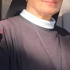 Uśmiechnięta siostra Bernadetta w czarnym habicie z białym kołnierzykiem, ozdobiona gałązką z różowymi kwiatami.