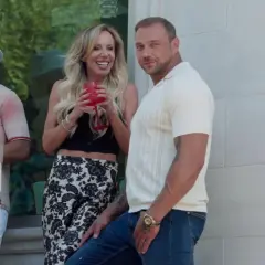 Dorota, żona dla polaka, śmieje się, trzymając drinka. Obok stoi mężczyzna w beżowej koszulce polo.