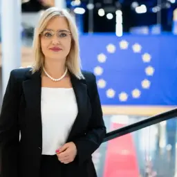 Magda Adamowicz, pogodynka, której życiorys jest pełen sukcesów, w eleganckim stroju na tle flagi UE.