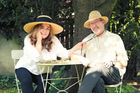 Para z kapeluszami siedzi przy stoliku w ogrodzie. Kobieta, jakby zainspirowana twórczością Daukszewicza, bawi się jego krawatem.