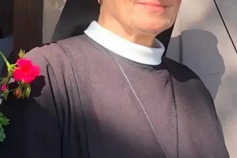 Uśmiechnięta siostra Bernadetta w czarnym habicie z białym kołnierzykiem, ozdobiona gałązką z różowymi kwiatami.