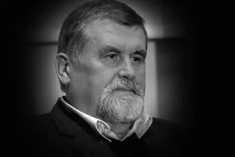 Portret Andrzeja Zielińskiego, mężczyzny z siwą brodą i zmarszczkami, w czarnym garniturze.
