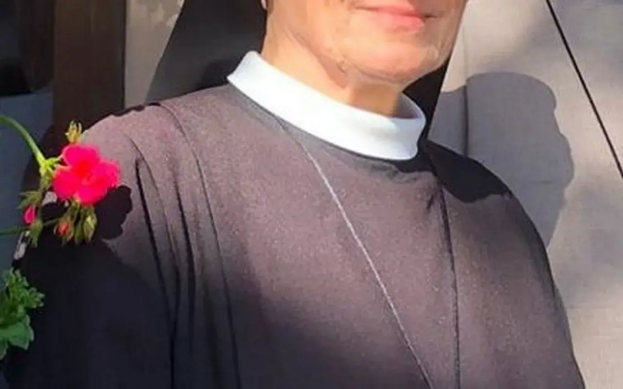 Uśmiechnięta siostra Bernadetta w czarnym habicie z białym kołnierzykiem, ozdobiona gałązką z różowymi kwiatami.