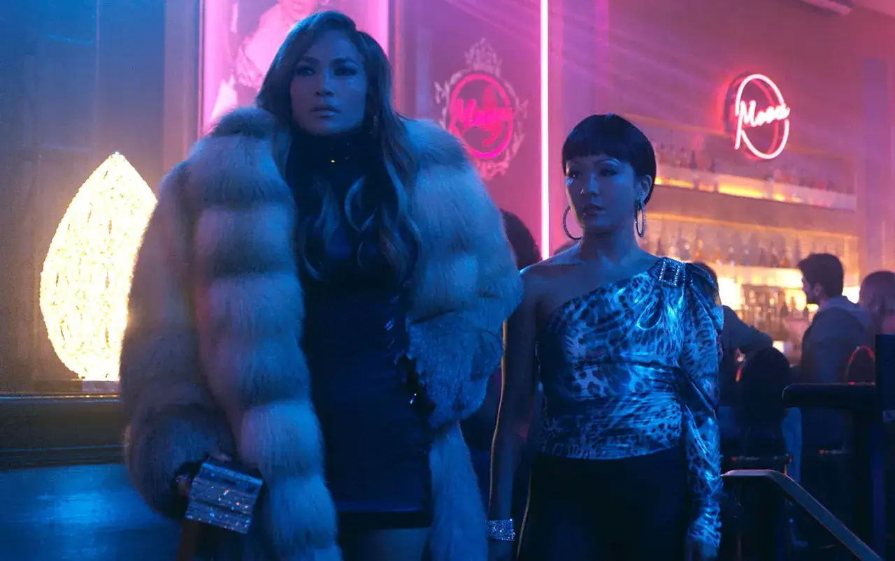 Jennifer Lopez w filmach, w neonowym klubie, w futrze i z błyszczącą kopertówką. Obok niej stoi kobieta w panterkowej bluzce.