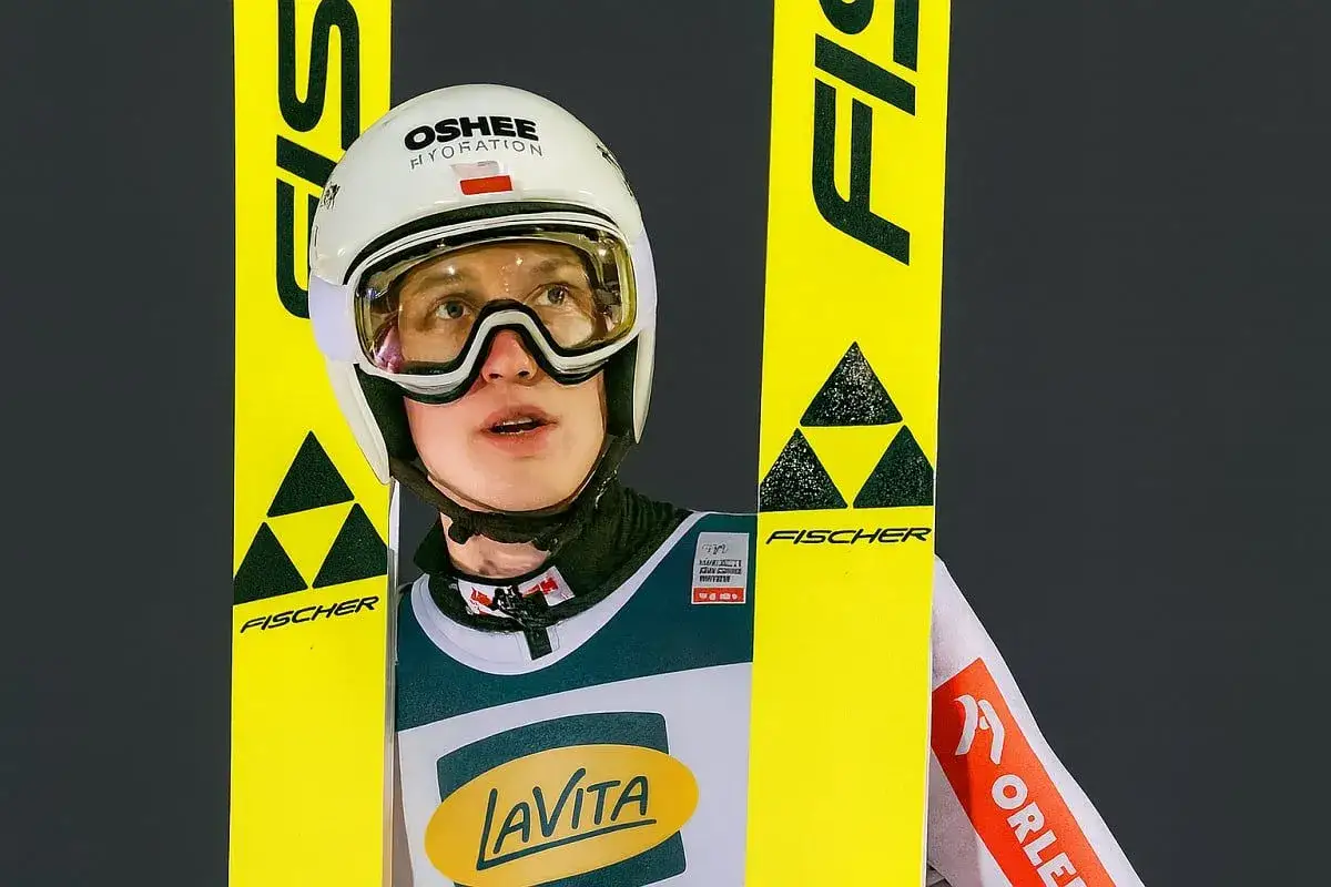 Kacper Tomasiak, skoczek narciarski, w kasku i goglach, gotowy do skoku. Obok niego żółte narty Fischer.