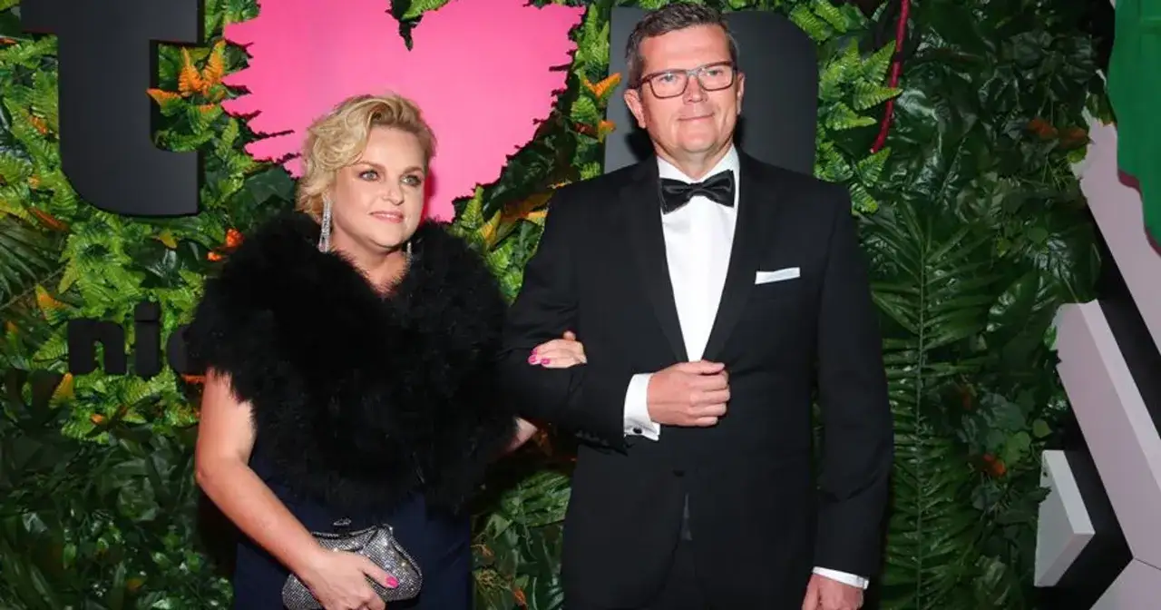 Marcin Bosacki w garniturze i jego partnerka w czarnym futrze, na tle zieleni i różowego serca.