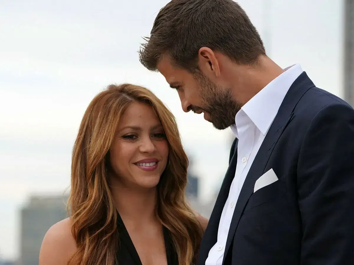 Shakira i Pique patrzą na siebie z uśmiechem. Ona ma długie, falowane włosy, on elegancki garnitur.
