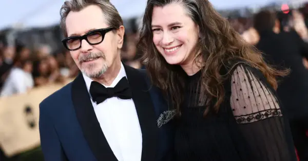 Gary Oldman z żoną, oboje uśmiechnięci, on w eleganckim garniturze, ona w czarnej sukni z koronkowymi rękawami.